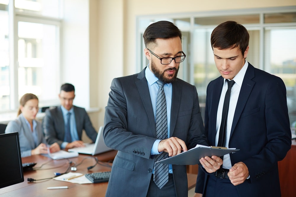Deux professionnels en costume discutent et consultent des documents dans une salle de réunion, illustration d’un audit ou vérification ISO. comment savoir si une entreprise est certifiée iso.